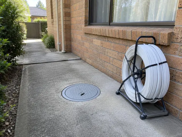 Concrete side path with a capped inspection cover beside a brick house and a pipe reel in St Ives