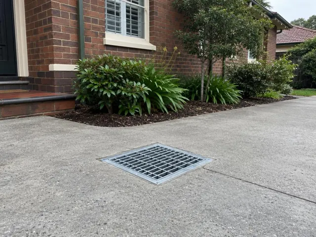 Concrete driveway with a seated stormwater grate beside a brick house and garden in Roseville
