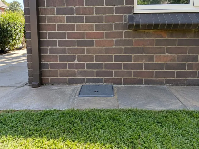 Flush capped sewer access opening in a concrete path beside a brick house wall in Gordon